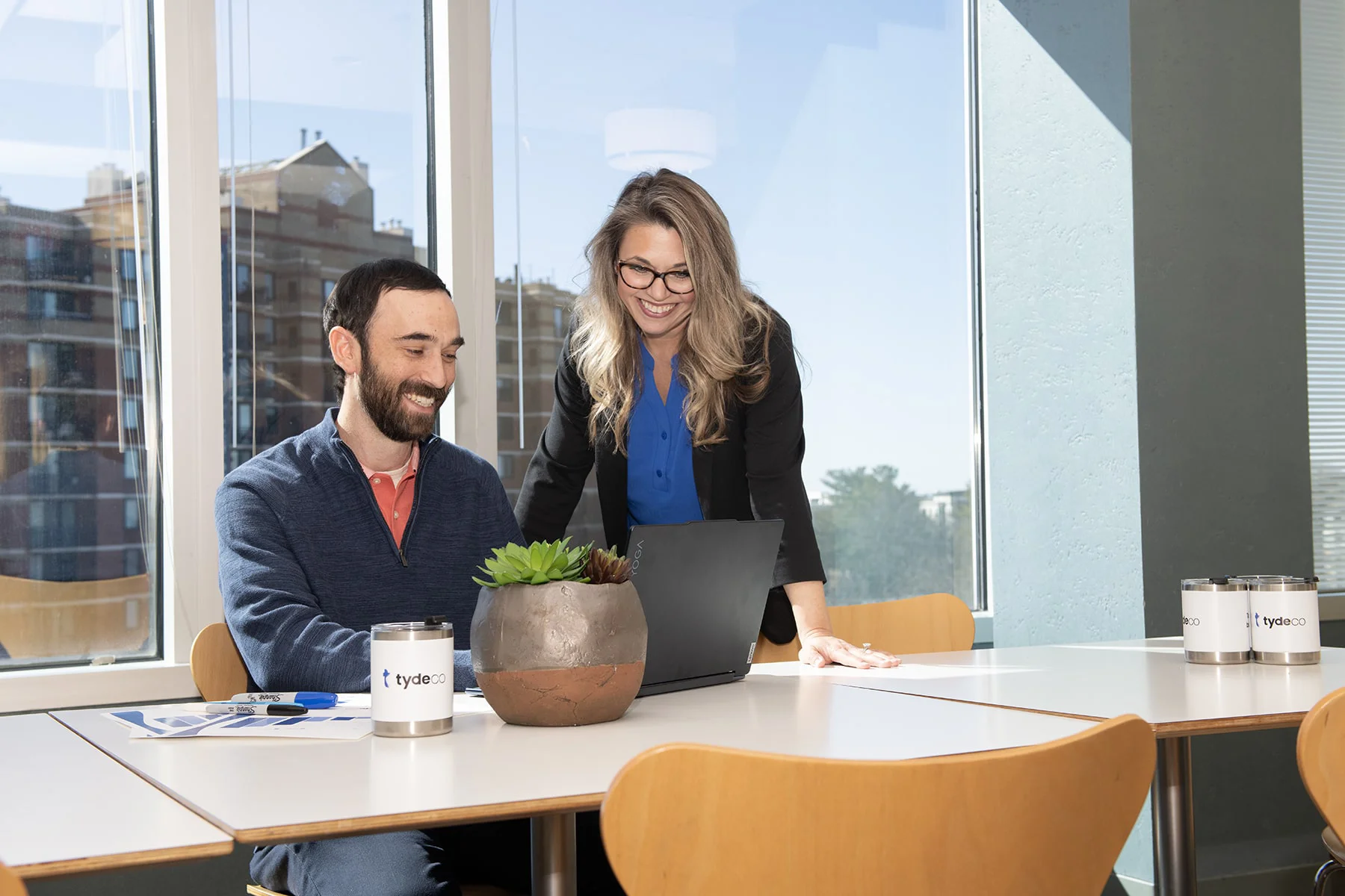 man and a woman looking at a laptop in an office with the view of a building in the background