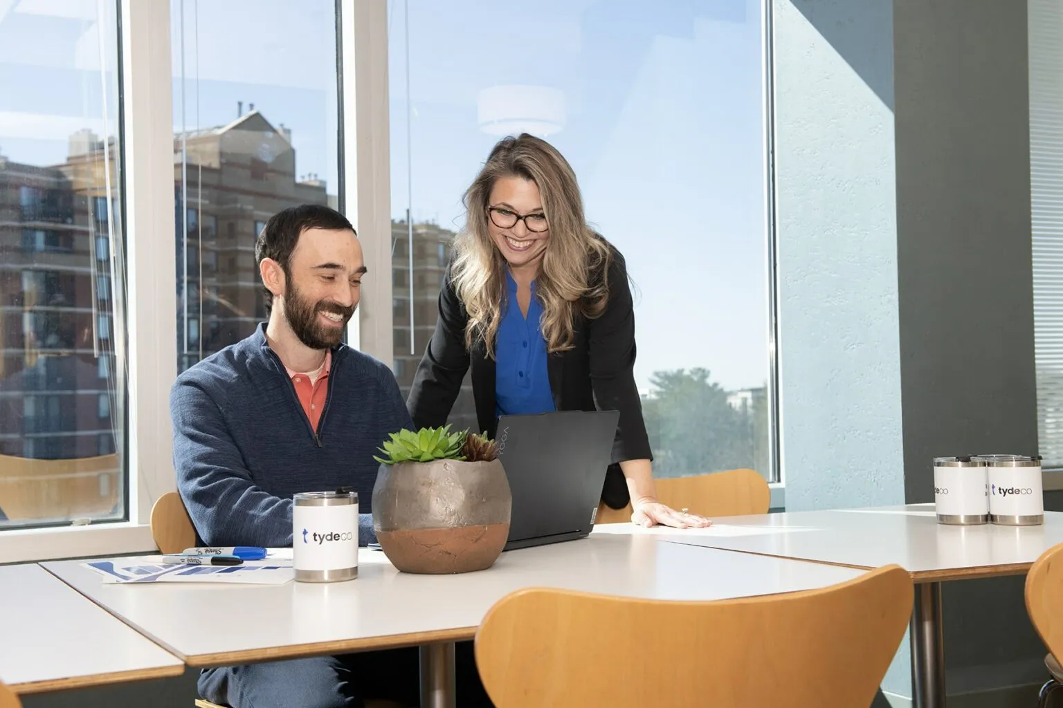 man and a woman looking at a laptop in an office with the view of a building in the background