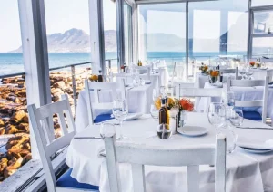 white table set for dining in a bright seaside restaurant with ocean and mountains visible through glass walls.