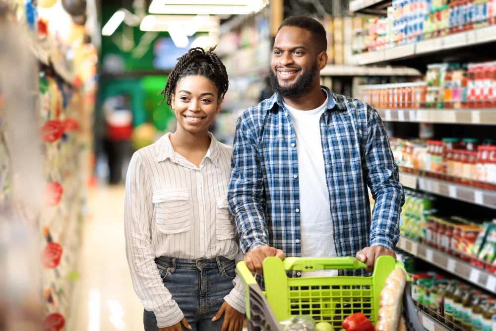 Smiling couple shopping together in a grocery store aisle with a cart filled with food items.