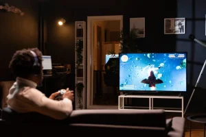 A man with headphones sits on a couch in a dark room, playing a space shooter video game on a large TV.
