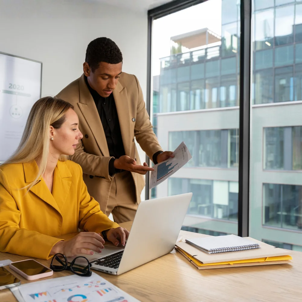 A woman in a yellow blazer sits at a desk using a laptop while a man in a tan suit stands beside her, holding a paper with a graph and gesturing with his hand. Documents, folders, and glasses are on the desk, and an office building is visible through a window behind them.