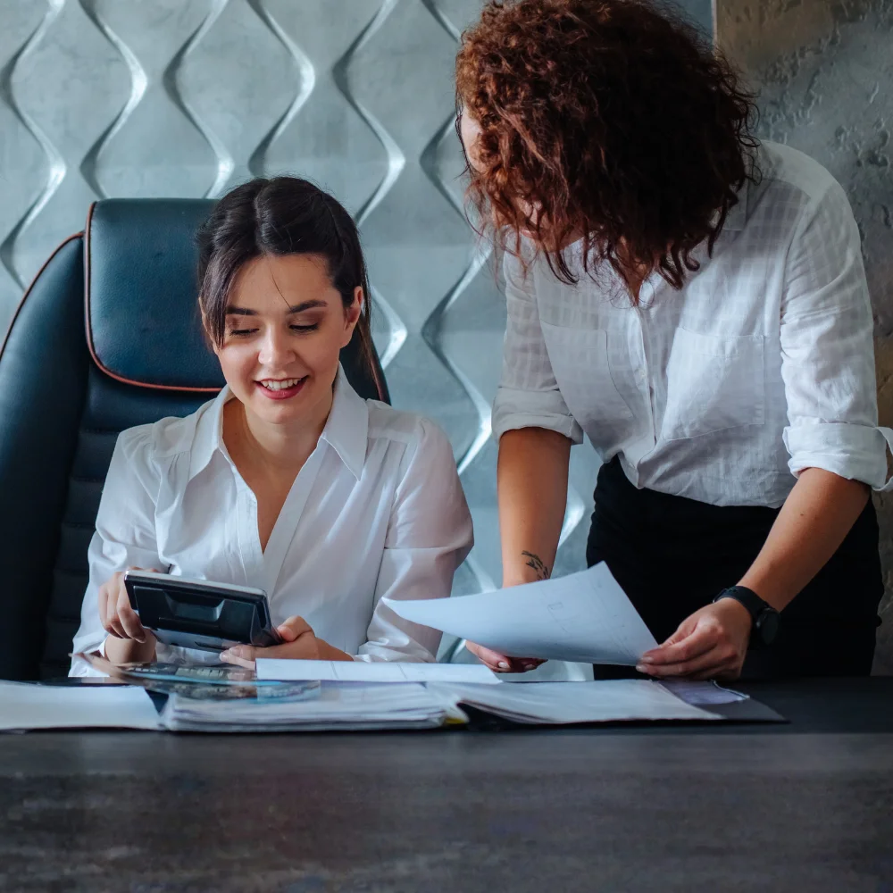 A female manager with curly reddish-brown hair stands at a desk in a modern office, showing documents to a seated smiling woman who holds a calculator and reviews files. Both women wear white button-down shirts.