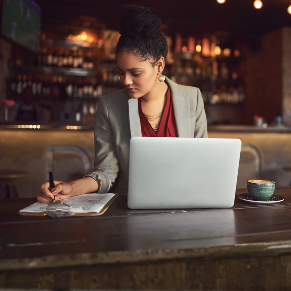 A woman in a grey blazer writes on a clipboard next to an open laptop and a cup of coffee at a bar top in a dimly lit restaurant. She has dark hair styled in a high bun.
