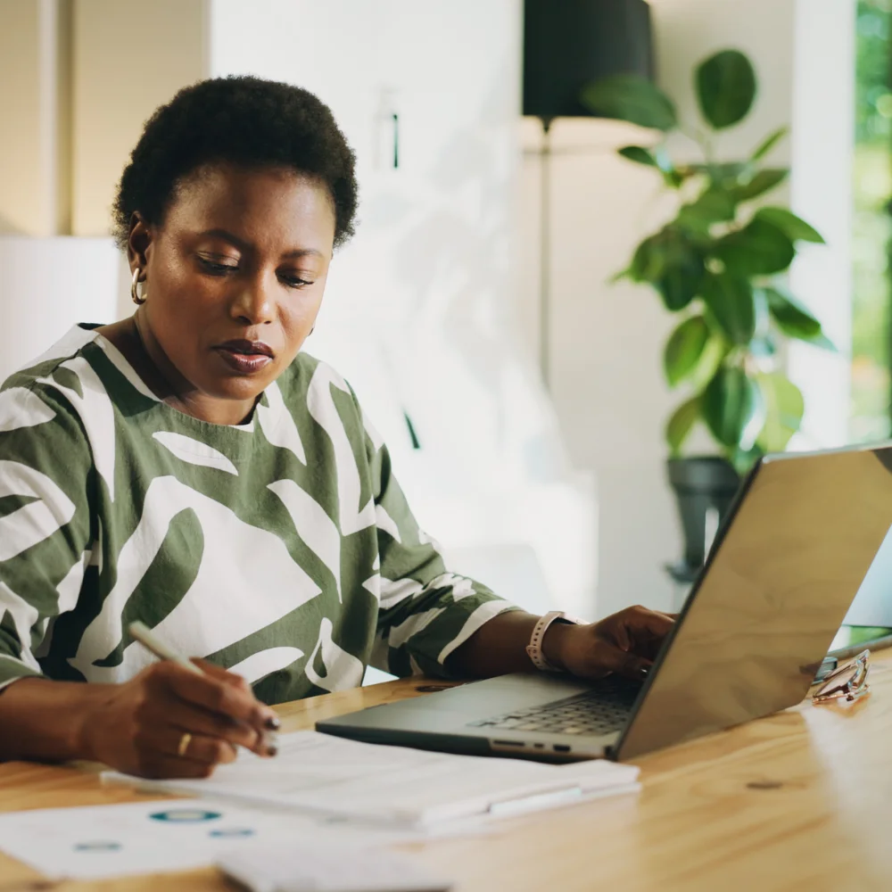 A medium close-up view capturing an African woman working at her home office table in the sunlight. In the image, she is sitting, looking down while taking notes on papers with a pen. An open laptop, glasses, and documents with charts sit on the wooden desk, and a potted plant and floor lamp decorate the background. The woman wears a patterned green and white shirt with three-quarter sleeves and a pink-strapped watch. This photo could serve as a stock photo to communicate a range of topics, from entrepreneurship and productivity to career and home offices.