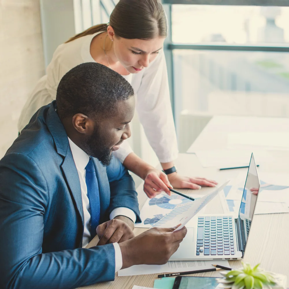 A medium-angle, full shot focuses on a female in a white long-sleeved shirt standing beside a seated male in a blue suit as they look down at a paper the male is holding. A blurred background with two windows suggests an office environment.