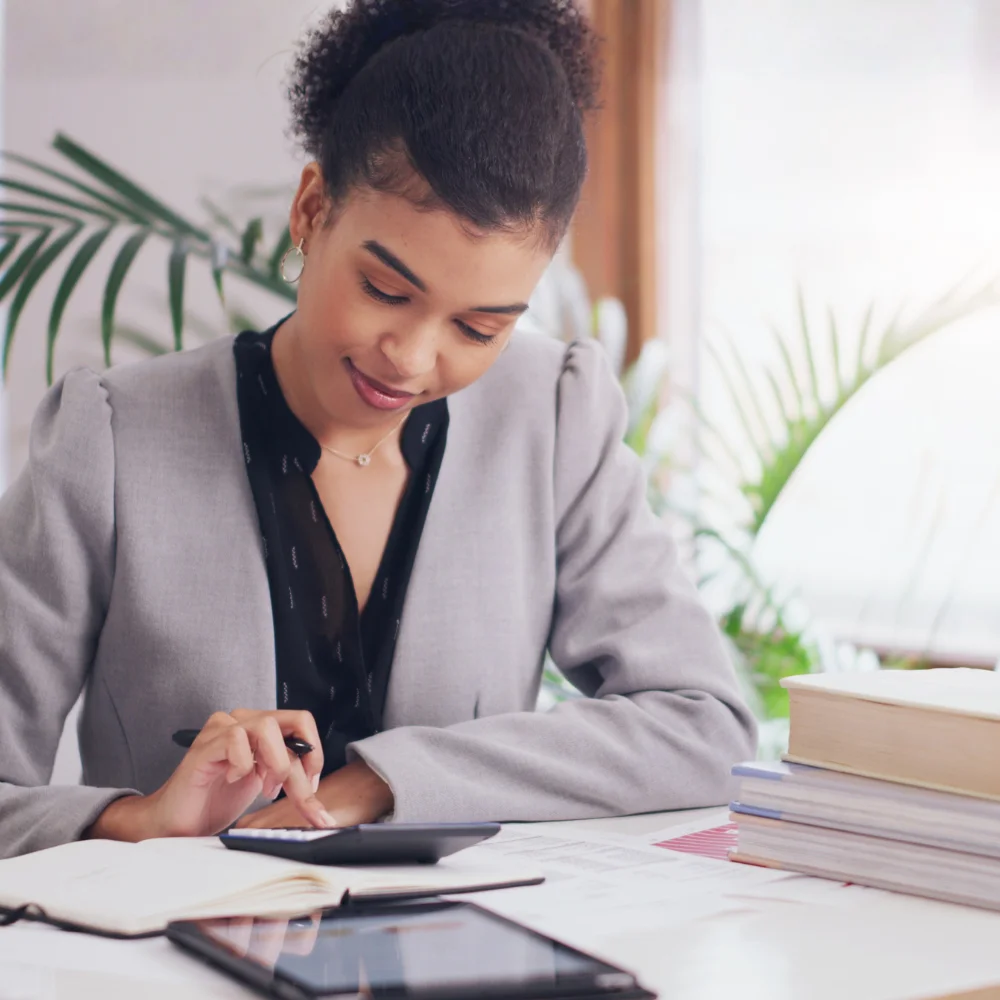 A medium-shot portrait depicts a Black woman with dark, curly hair tied back wearing a grey blazer over a black top smiling and looking down while typing on a calculator. Her right hand holds a pen while a small black calculator is under her left hand on top of an open notebook. Books, an out-of-focus tablet and plant life sit around her on a light-colored table in front of a brightly lit window.