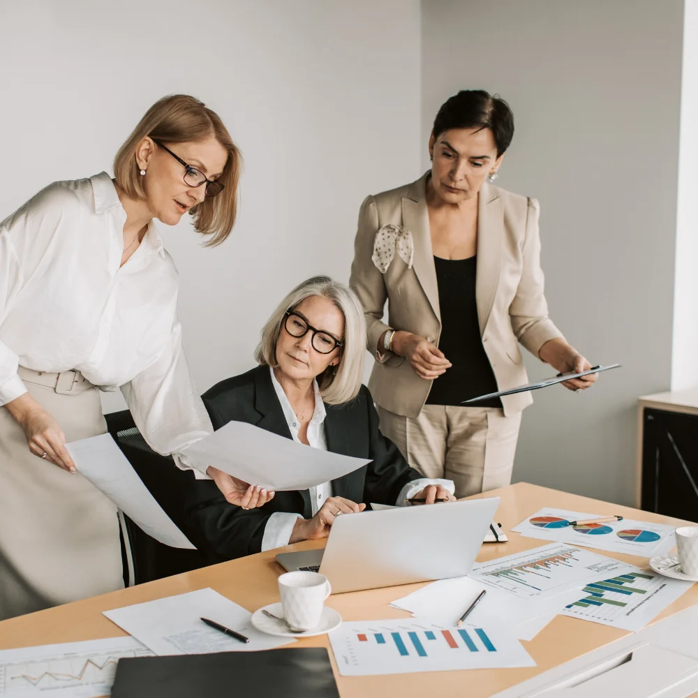 A medium-angle, full shot depicts three middle-aged women discussing papers and data in a well-lit modern office space. One woman with short blonde hair and reading glasses, wearing a white button-down and a cream skirt, points at and looks down at the data in the open notebook. Another woman wearing a matching beige pantsuit with black-framed glasses holding papers looks over at the woman in the pantsuit on her right. A third woman in a black blazer and white top, with shoulder-length grey hair and black-rimmed glasses, sits in front of a silver laptop and also looks towards the woman in the pantsuit. She holds papers in her right hand as her left hand rests on the laptop keyboard. On the light brown desk, four pieces of paper with bar charts and pie graphs are spread in front of the laptop, with two grey coffee cups on saucers.