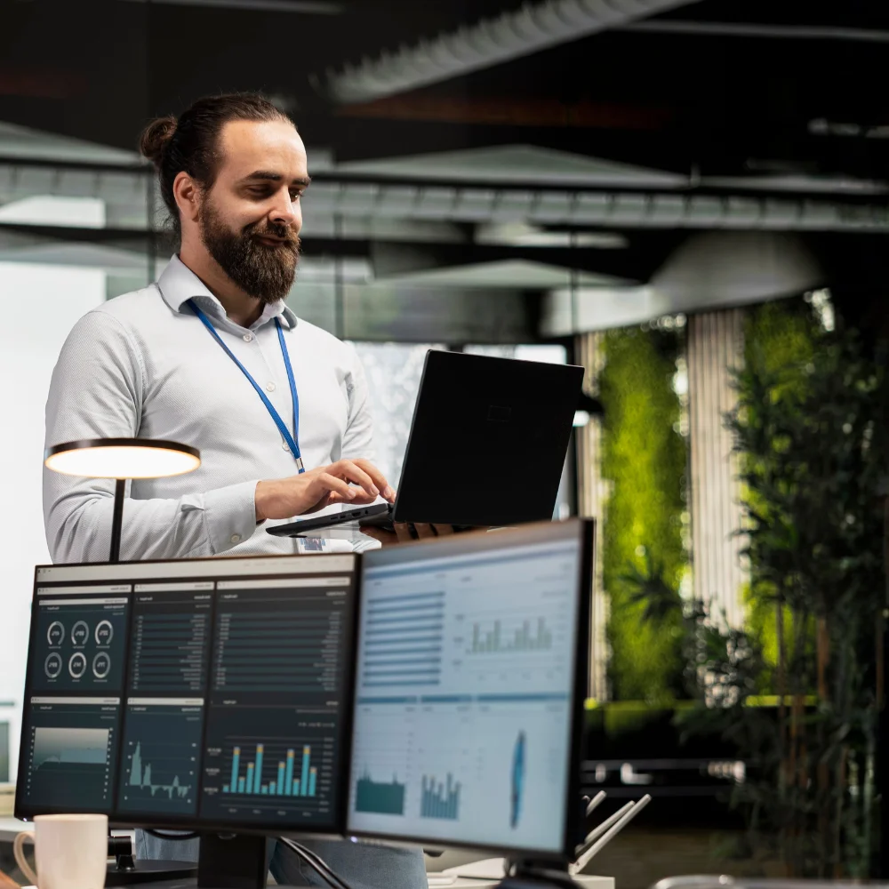 A medium shot shows a bearded man in a bun, white shirt, and a blue lanyard, looking at a black laptop he holds while working at a desk with two monitors displaying bar graphs, a desk lamp, and a mug. In the background is an open-plan office with green plants, large windows, and an industrial-style ceiling with dark pipes. He seems to be in the middle of a project or task and has a concentrated but relaxed expression on his face.