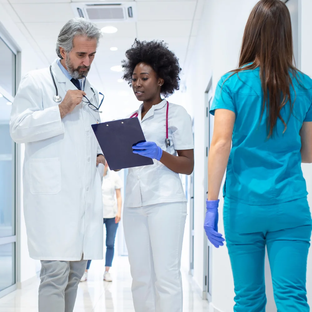 A busy hospital corridor where medical professionals are collaborating. A senior male doctor in a white lab coat and stethoscope listens intently to a female nurse with a clipboard, while another healthcare worker in blue scrubs walks by in the foreground. The bright, sterile environment of the medical center highlights a scene of professional teamwork and patient care management.