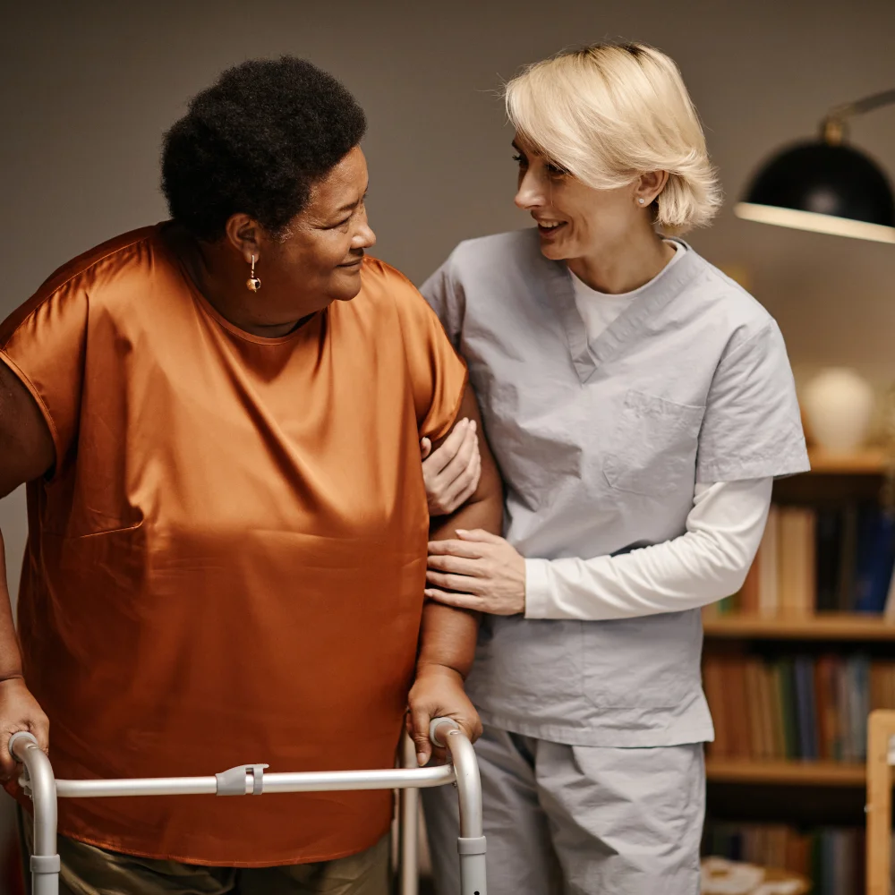 A heartwarming scene of long-term care featuring a blonde female caregiver in light blue scrubs supporting an elderly Black woman. The caregiver gently holds the woman’s arm as she uses a silver walker, and they share a warm, smiling glance. The cozy, dimly lit indoor setting with a bookshelf in the background conveys a sense of safety, dignity, and personalized support in a residential care facility.