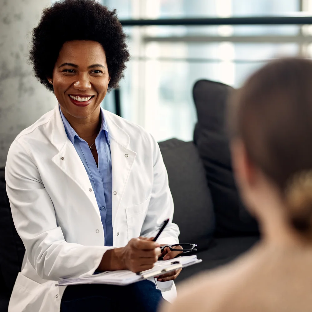 A warm and reassuring primary care consultation. A female doctor with a kind smile, wearing a white lab coat over a blue shirt, sits with a clipboard and pen while looking at a patient (seen from the back). The bright, modern indoor setting and the doctor's attentive expression emphasize a professional yet compassionate healthcare experience.